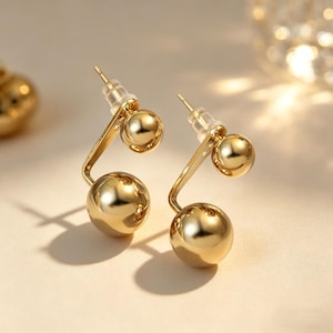 May include: A pair of gold-colored, double-sided ball earrings. Each earring features a small ball on the front and a larger ball on the back, connected by a curved gold bar. The earrings have a shiny, reflective surface.