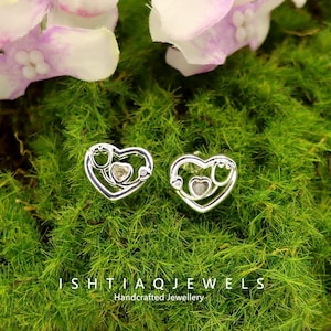 May include: A pair of silver heart-shaped earrings with a stethoscope design. Each earring has a small heart-shaped stone in the center. The earrings are displayed on a bed of green moss, with pink and white flowers in the background. The text "ISHTIAQ JEWELS Handcrafted Jewellery" is at the bottom.