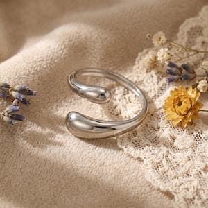 May include: A silver-colored, open-ended ring with a smooth, teardrop-shaped design. The ring curves around, with the teardrop shapes at each end. The ring is displayed on a beige surface with dried flowers and lace.