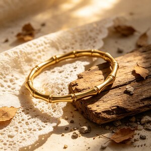 May include: A gold-colored bamboo-style bracelet. The bracelet is made of a shiny metal and is resting on a piece of wood. The background includes a white lace cloth and scattered dried leaves.