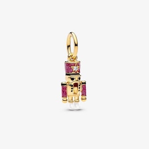 May include: A gold-toned nutcracker charm featuring a glittery magenta hat and sleeves, a small clear stone set in a star on the hat, black painted details on the face and uniform, and white painted boots, suspended from a circular gold jump ring.