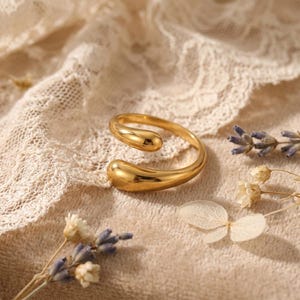 Könnte beinhalten: Ein goldfarbener Ring mit einem einzigartigen, offenen Design. Der Ring hat zwei tropfenförmige Enden, die sich umeinander krümmen. Er wird auf einem beigefarbenen Spitzengewebe mit getrockneten Blumen und Lavendelzweigen im Hintergrund präsentiert.