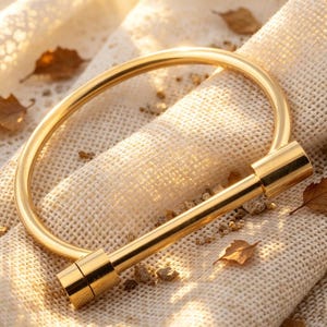 May include: A gold-colored bangle bracelet with a cylindrical bar closure. The bracelet is set against a textured, off-white fabric, with scattered dried leaves and small pebbles. The bracelet's design is simple and elegant.