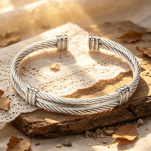 Puede incluir: Una pulsera de cable plateada con un diseño tejido. La pulsera presenta un patrón de cable grueso y retorcido con tapas de extremo lisas y redondeadas. La pulsera se exhibe sobre una superficie de madera con un mantel de encaje y hojas de otoño.