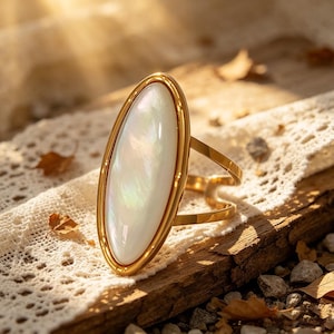 Op de afbeelding: Een goudkleurige ring met een grote, ovale, iriserende witte steen. De steen is gezet in een gouden frame en de ring heeft een eenvoudige, elegante band. De ring wordt gepresenteerd op een houten oppervlak met kant en gevallen bladeren.
