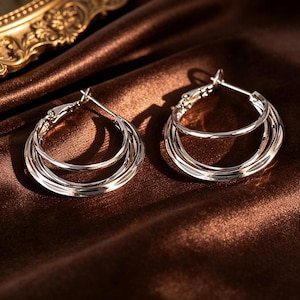 May include: Silver hoop earrings with a layered design. Each earring has three interconnected hoops, creating a multi-dimensional effect. The earrings are displayed on a brown satin surface, reflecting light and adding elegance.