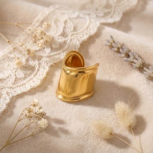 Op de afbeelding: Een goudkleurige ring met een brede, gebogen band. De ring heeft een glad, gepolijst oppervlak en een modern design. De ring wordt gepresenteerd op een crèmekleurig oppervlak met gedroogde bloemen en kant.