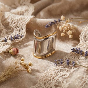 May include: A silver cuff bracelet with gold edging. The bracelet has a rectangular shape with a curved top and bottom. The background includes dried flowers and lace.