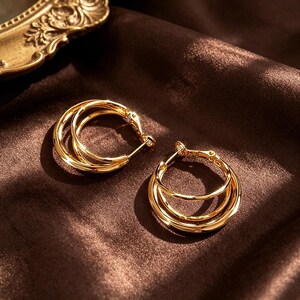 May include: A pair of gold-colored hoop earrings. Each earring features a triple-layered design, with three concentric hoops. The earrings are displayed on a dark brown, satin-like fabric, creating a contrast with the shiny gold.