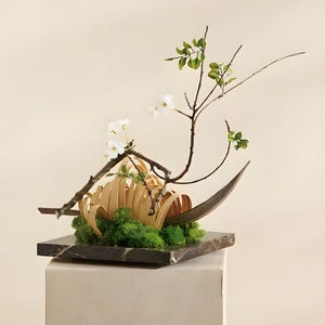 May include: A floral arrangement featuring white blossoms on thin brown branches, curved light-colored wooden strips, and green moss. The arrangement is set on a square, dark gray marble base with white veining, creating a structured display.