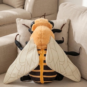 Może przedstawiać: Pluszowy plecak w kształcie pszczoły z żółto-czarnym paskowanym korpusem, beżowymi skrzydłami i żółtym zamkiem błyskawicznym. Pszczoła ma czarne czułki i nogi. Plecak spoczywa na beżowej poduszce i beżowej sofie.