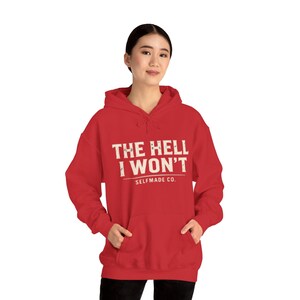 May include: A red hooded sweatshirt with the text "THE HELL I WON'T" and "SELFMADE CO." printed in white. The sweatshirt has a front pocket and a drawstring hood. The model is wearing the sweatshirt.