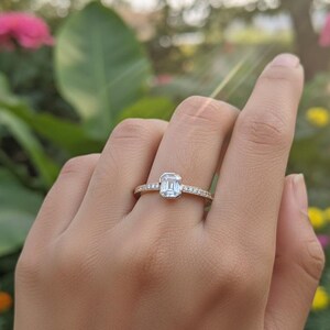 May include: A gold ring with a rectangular, light blue gemstone at the center. The band is adorned with small, clear gemstones. The ring is on a person's finger, with a blurred background of green foliage and flowers.
