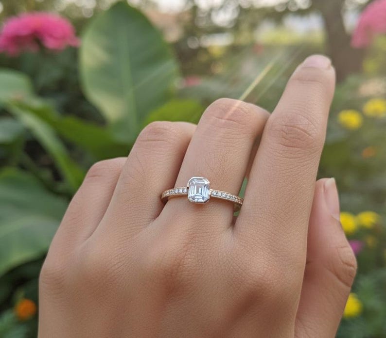 May include: A gold ring with a rectangular, light blue gemstone at the center. The band is adorned with small, clear gemstones. The ring is on a person's finger, with a blurred background of green foliage and flowers.