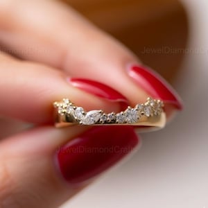 Può includere: Un anello a fascia in oro con un design curvo, incastonato con più pietre preziose trasparenti. L'anello presenta un mix di pietre rotonde e a taglio marquise, creando un aspetto scintillante ed elegante. L'anello è tenuto da una persona con lo smalto rosso.
