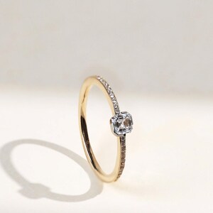 May include: A gold ring with a square, clear gemstone at the center. The band is thin, with small, clear stones set along one side. The ring is set against a soft, white background, with a shadow cast to the left.