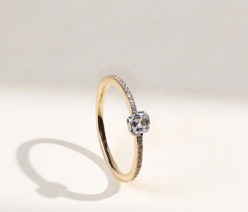 May include: A gold ring with a square, clear gemstone at the center. The band is thin, with small, clear stones set along one side. The ring is set against a soft, white background, with a shadow cast to the left.