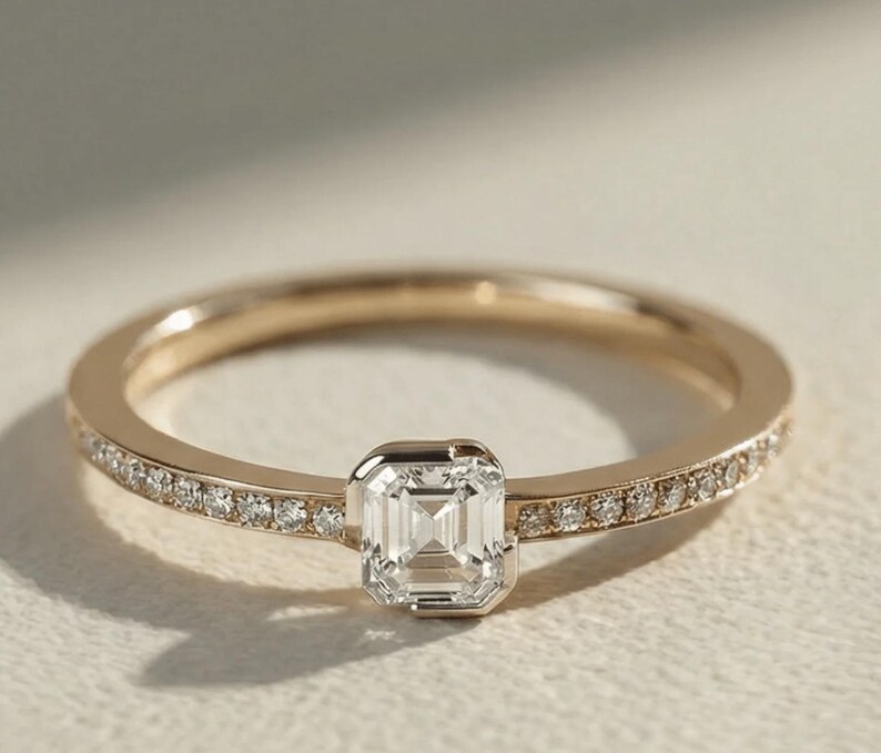 May include: A gold ring with a square-cut, clear gemstone at the center. The band is lined with small, clear gemstones. The ring is set against a soft, neutral background, highlighting its elegant design.