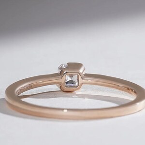 May include: Rose gold ring with a square-cut, clear gemstone. The ring band is thin and polished, with a subtle inscription. The gemstone is set in a square bezel, creating a minimalist and elegant design. The ring is shown on a white surface.