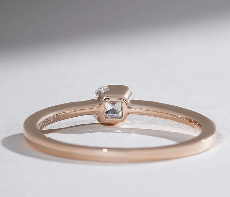 May include: Rose gold ring with a square-cut, clear gemstone. The ring band is thin and polished, with a subtle inscription. The gemstone is set in a square bezel, creating a minimalist and elegant design. The ring is shown on a white surface.