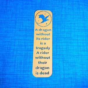 May include: A tan bookmark with a dragon silhouette and the text "A dragon without its rider is a tragedy. A rider without their dragon is dead." The bookmark is on a blue textured surface.