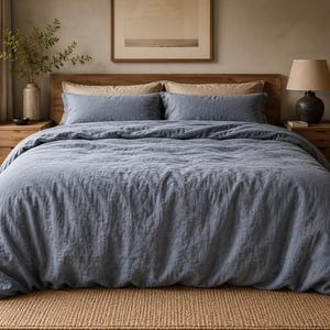 Peut inclure: Un lit doté d'une housse de couette en lin bleu et de deux taies d'oreiller rectangulaires assorties. La literie est disposée devant une tête de lit en bois avec deux tables de chevet en bois de chaque côté, chacune surmontée d'une lampe.