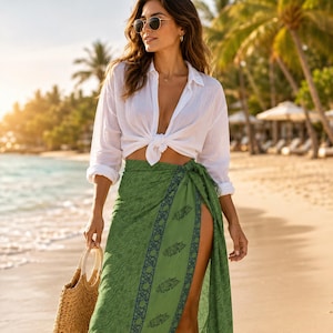Puede incluir: Una mujer con una camisa blanca anudada a la cintura y una falda pareo verde con estampado floral. Lleva un bolso tote tejido y gafas de sol. El fondo muestra una playa con palmeras y sombrillas.