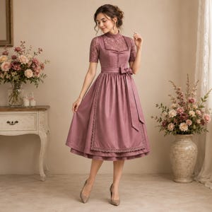May include: A dusty rose midi-length dirndl dress featuring a high neckline, short lace sleeves, and a structured bodice. The skirt includes a matching apron with delicate lace trim along the edges and a tied bow at the waist, creating a classic silhouette.