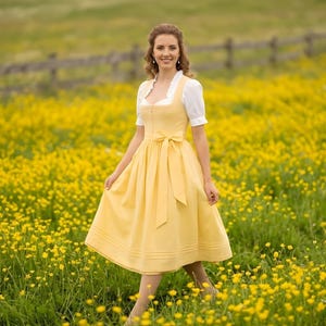 May include: A yellow dirndl dress with a white blouse and a bow at the waist. The dress has a knee-length skirt and is worn in a field of yellow flowers. The sleeves of the blouse are short and puffy.