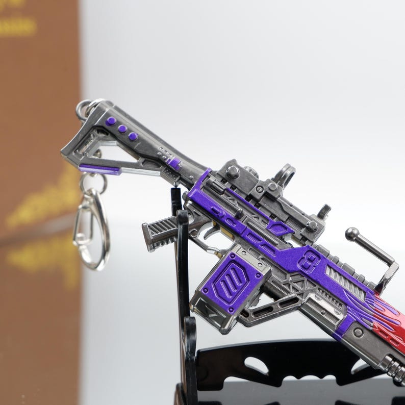 Puede incluir: Una pistola en miniatura detallada con un acabado morado y plateado. La pistola de juguete presenta detalles intrincados, que incluyen una mira, un asa y un cargador. El ca&ntilde;&oacute;n tiene un dise&ntilde;o de llama roja. Una llavero est&aacute; adjunto a la pistola.