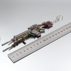Puede incluir: Un llavero de rifle en miniatura en tonos verdes, marrones y plateados. El llavero tiene un visor y est&aacute; unido a un anillo plateado. Una regla met&aacute;lica con marcas de cent&iacute;metros est&aacute; al lado.