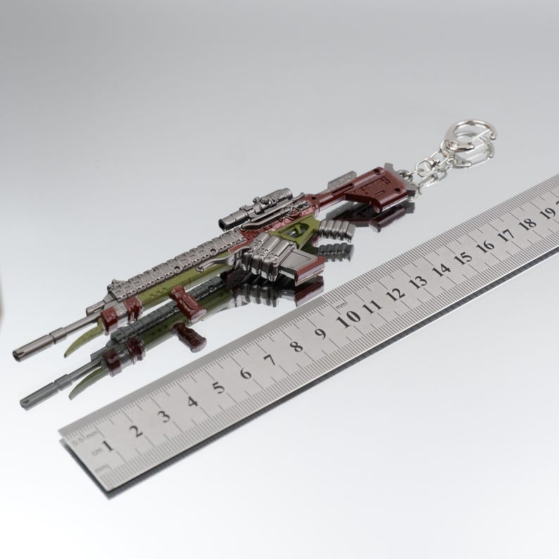 Puede incluir: Un llavero de rifle en miniatura en tonos verdes, marrones y plateados. El llavero tiene un visor y est&aacute; unido a un anillo plateado. Una regla met&aacute;lica con marcas de cent&iacute;metros est&aacute; al lado.