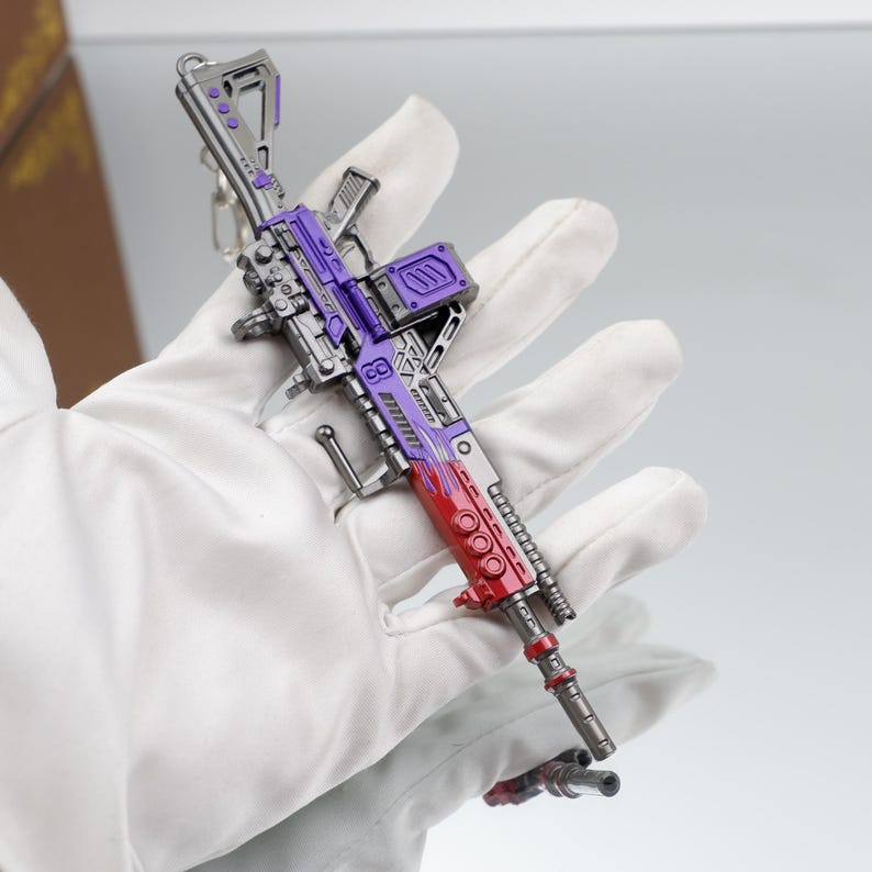 Puede incluir: Un juguete en miniatura con forma de pistola, en plata, morado y rojo. El juguete se sostiene en una mano enguantada, destacando su dise&ntilde;o detallado. Tiene un llavero. El dise&ntilde;o es intrincado y el tama&ntilde;o es peque&ntilde;o.