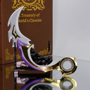 Puede incluir: Una hoja curva decorativa con una hoja blanca y morada, detalles dorados y un mango circular. La hoja se exhibe sobre un soporte negro, con un libro marrón en el fondo que dice "A Treasury of World's Classies."