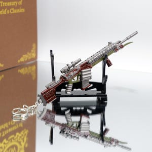 Puede incluir: Un llavero de rifle en miniatura con mira telesc&oacute;pica, en tonos plateados, rojos y verdes. El arma de juguete est&aacute; montada sobre un soporte negro, con una superficie reflectante debajo. El llavero est&aacute; sujeto con una cadena y un anillo plateados.