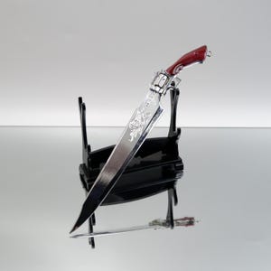 Puede incluir: Un cuchillo decorativo plateado y rojo, presentado sobre un soporte negro. La hoja presenta un diseño grabado. El mango es de color rojo oscuro. El cuchillo es un objeto de colección.