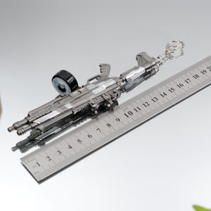 Puede incluir: Un llavero de rifle en miniatura de color plateado con un cargador negro. El llavero mide aproximadamente 18 cm de largo y presenta detalles intrincados. Incluye un llavero para sujetarlo. El art&iacute;culo se muestra sobre una superficie reflectante.