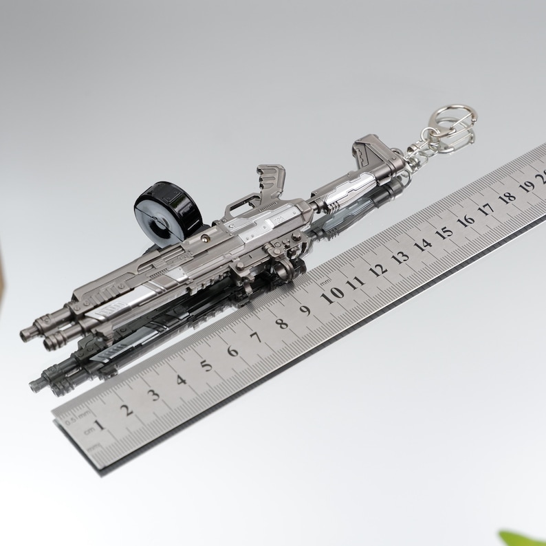 Puede incluir: Un llavero de rifle en miniatura de color plateado con un cargador negro. El llavero mide aproximadamente 18 cm de largo y presenta detalles intrincados. Incluye un llavero para sujetarlo. El art&iacute;culo se muestra sobre una superficie reflectante.