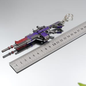 Puede incluir: Un llavero en miniatura con forma de pistola, en plata, morado y rojo. El llavero mide aproximadamente 18 cm de largo e incluye un llavero de metal. El dise&ntilde;o presenta detalles intrincados, incluyendo una mira y varios componentes mec&aacute;nicos.