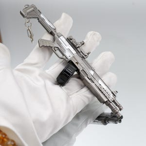 Puede incluir: Un rifle en miniatura plateado y negro con un llavero adjunto. El juguete tiene un cargador, mira telesc&oacute;pica y un dise&ntilde;o detallado. El objeto se sostiene en una mano enguantada.