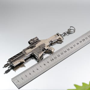 Puede incluir: Un llavero detallado en forma de pistola met&aacute;lica con mira y dise&ntilde;o intrincado. El llavero mide aproximadamente 20 cm de largo, con un llavero plateado. El dise&ntilde;o incluye una regla para la escala.