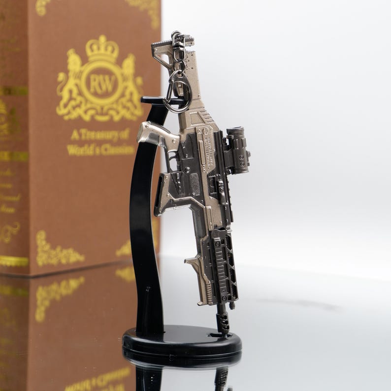 Puede incluir: Un llavero de rifle en miniatura plateado y negro con mira telesc&oacute;pica y detalles intrincados. El llavero est&aacute; montado en un soporte negro. Un libro marr&oacute;n con letras doradas se ve en el fondo.