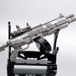 Puede incluir: Una detallada pistola de juguete gris met&aacute;lico con un cargador de tambor negro y una mira telesc&oacute;pica. La pistola est&aacute; montada en un soporte de exhibici&oacute;n negro. El dise&ntilde;o incluye detalles intrincados y una est&eacute;tica futurista.