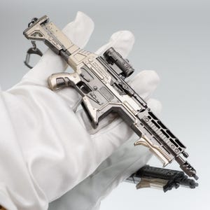 Puede incluir: Un rifle de asalto en miniatura de color plateado con mira telesc&oacute;pica y cargador desmontable. El rifle tiene detalles intrincados y se sostiene en una mano enguantada. El texto "HANUDO ASSAULT RIFLE ASSAULT RIFLE" es visible en el lateral.