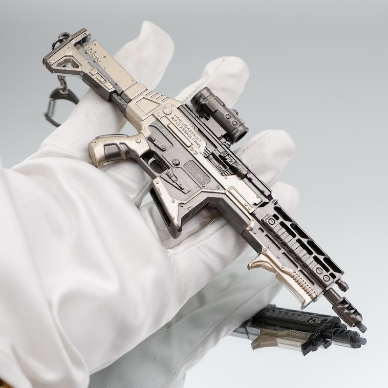 Puede incluir: Un rifle de asalto en miniatura de color plateado con mira telesc&oacute;pica y cargador desmontable. El rifle tiene detalles intrincados y se sostiene en una mano enguantada. El texto "HANUDO ASSAULT RIFLE ASSAULT RIFLE" es visible en el lateral.