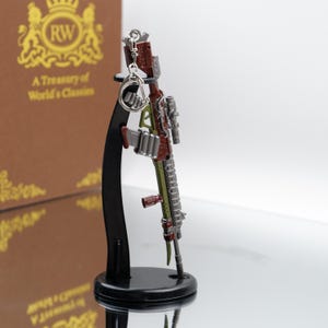 Puede incluir: Un llavero de rifle en miniatura detallado en tonos rojos, verdes y plateados, montado en un soporte de exhibici&oacute;n negro. El llavero presenta detalles intrincados y un dise&ntilde;o realista.