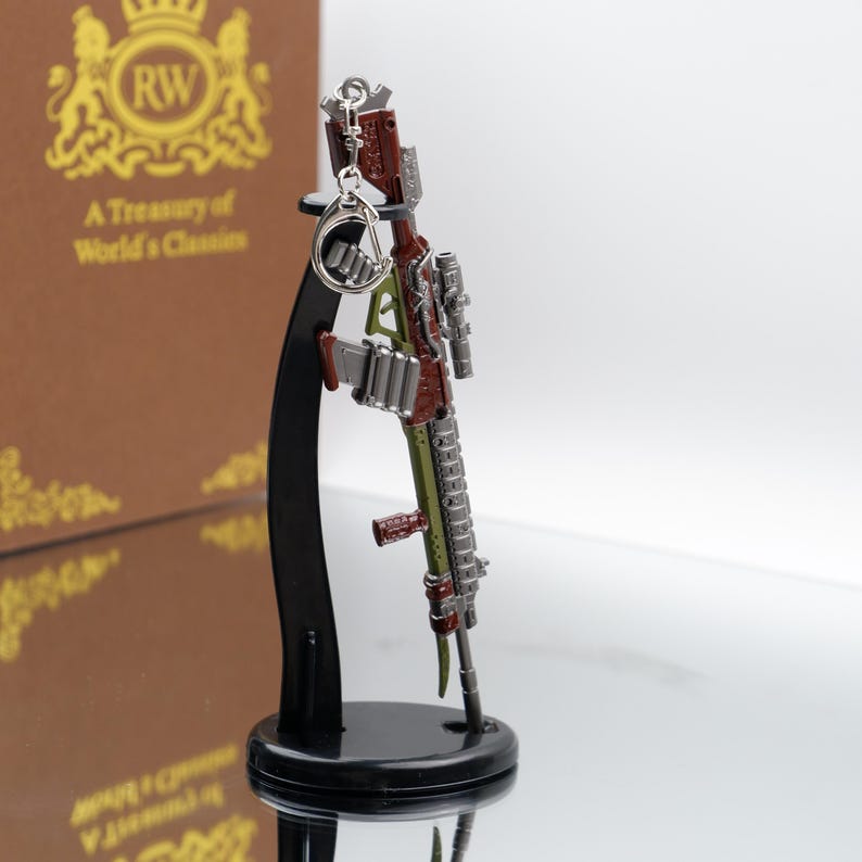 Puede incluir: Un llavero de rifle en miniatura detallado en tonos rojos, verdes y plateados, montado en un soporte de exhibici&oacute;n negro. El llavero presenta detalles intrincados y un dise&ntilde;o realista.