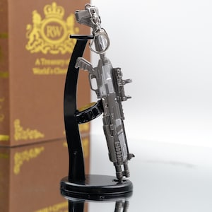 Puede incluir: Un llavero de rifle en miniatura de color plateado con detalles intrincados, que incluyen una mira telesc&oacute;pica y un cargador. El llavero est&aacute; unido a una peque&ntilde;a pistola plateada. Se exhibe en un soporte negro con una base circular.