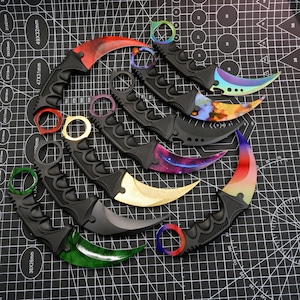 Peut inclure: Collection de couteaux karambit avec lames incurvées et anneaux pour les doigts. Les lames présentent diverses couleurs et motifs, notamment des motifs rouges, dorés et galactiques. Les manches sont noirs avec des poignées texturées.