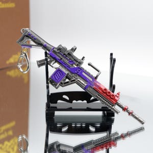 Puede incluir: Un rifle en miniatura con un esquema de color p&uacute;rpura y rojo, con una mira telesc&oacute;pica y un llavero. El arma de juguete se exhibe sobre un soporte negro, con una superficie reflectante debajo.